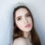 A bride with long brown hair, wearing a tiara and veil, looking at the camera.