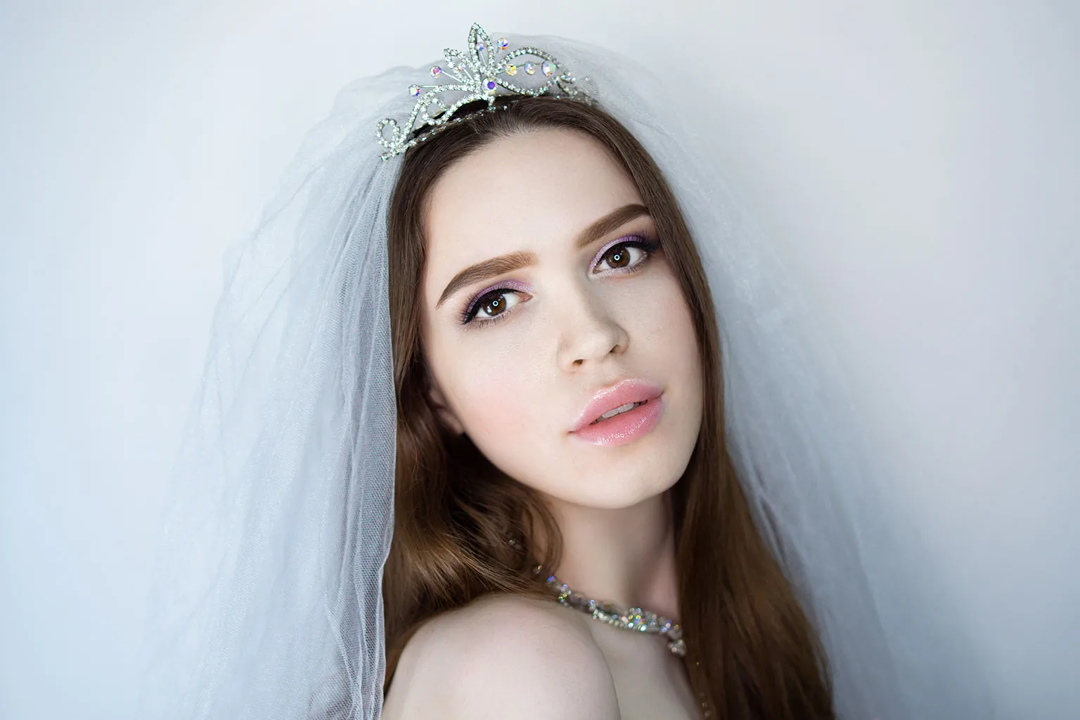 A bride with long brown hair, wearing a tiara and veil, looking at the camera.