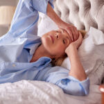 Woman lying in bed touching her forehead, looking unwell.